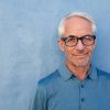 Close up portrait of handsome senior man with glasses looking at camera, he is standing against a blue wall.